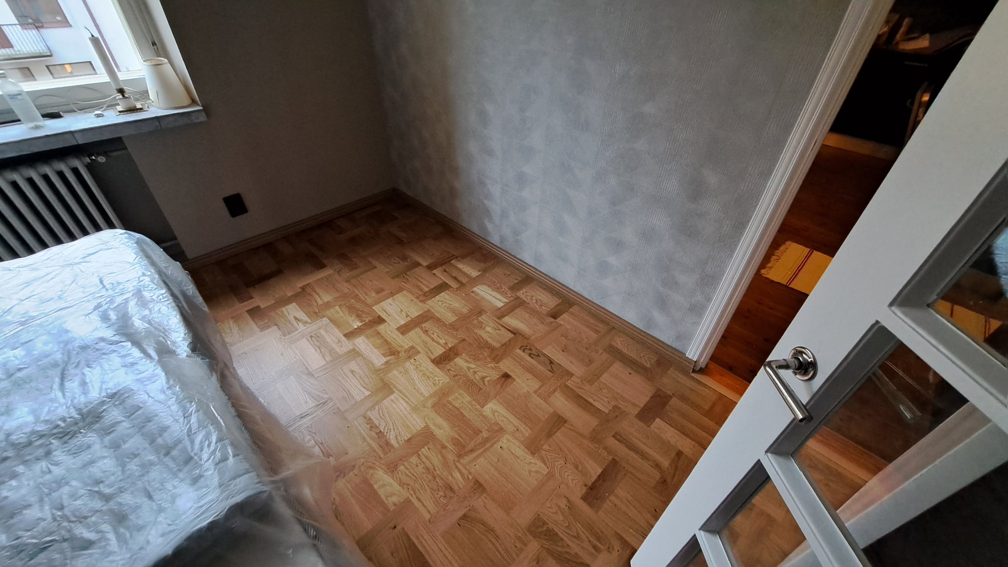 Parkettilattian korjaus ja asennus huoneessa, jossa on vaalea puupintainen mosaiikkiparketti ja valkoiset lasiovet.
