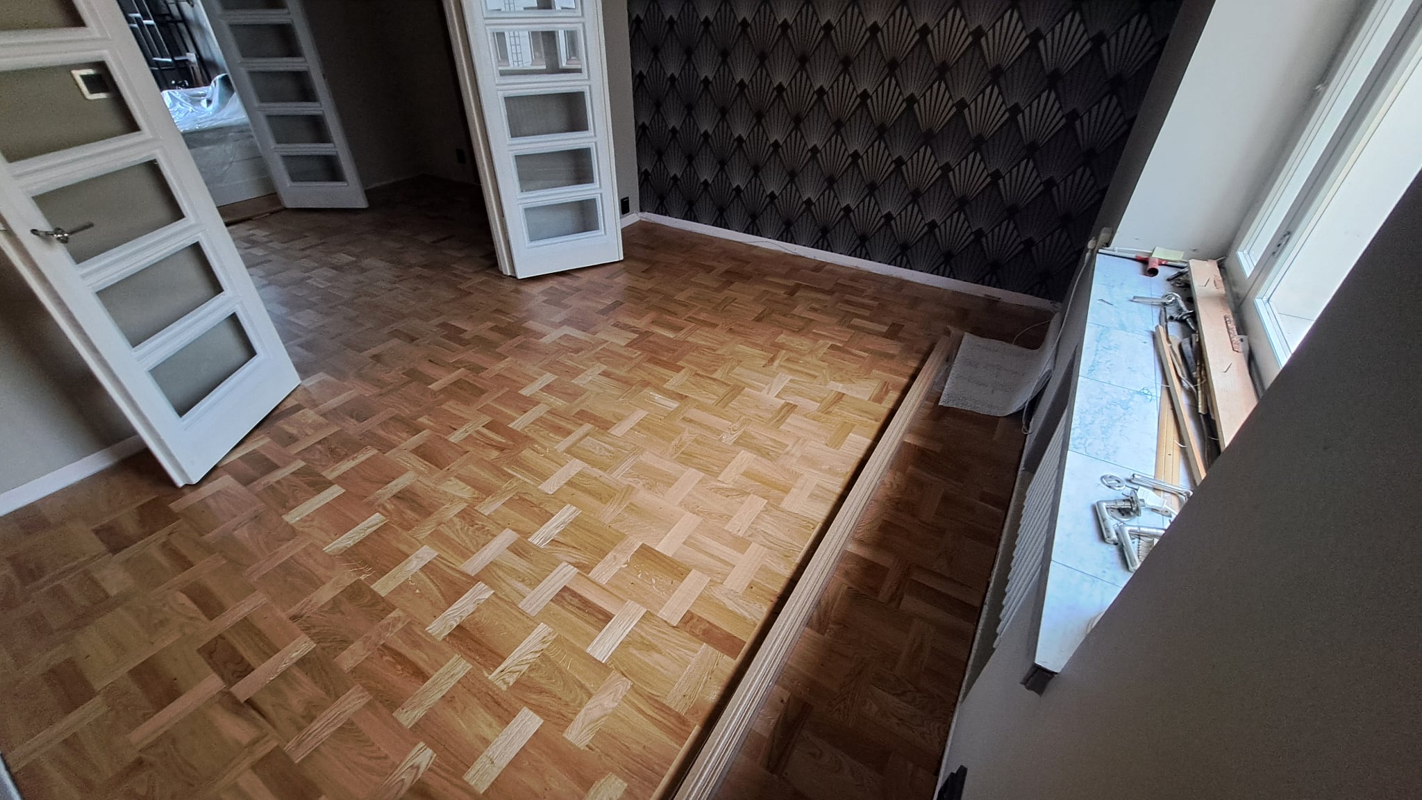 Mosaiikkiparketin kunnostus ja lattian viimeistely huoneessa, jossa on tummat tapetoidut seinät ja valkoiset lasiovet.